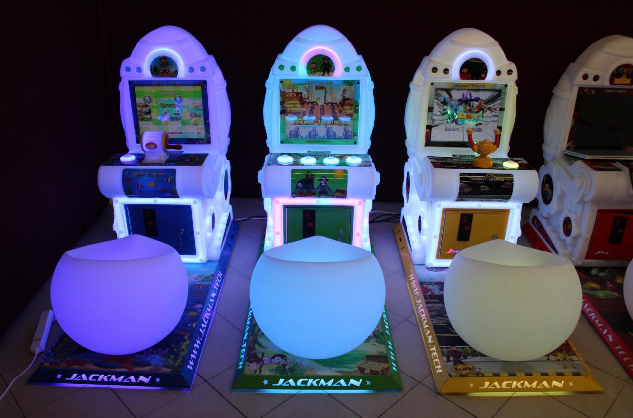 Trzy podświetlane automaty do gier o futurystycznym wyglądzie, z okrągłymi podświetlanymi siedziskami przed każdym z nich, każdy automat z inną grą na ekranie i innym kolorem podświetlenia.