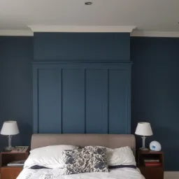 Pomieszczenie sypialne z łóżkiem, szafkami nocnymi i lampkami, ściana w odcieniach granatu z dekoracyjną zabudową, białe listwy przypodłogowe i sufitowe, zasłona w oknie.