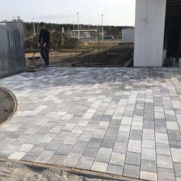 Denysov - Prace brukarskie w toku: zagęszczarka ubija szaro-białą kostkę brukową ułożoną we wzór, widoczne obrzeża i przygotowane podłoże.