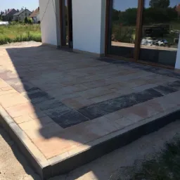 Wykończony taras z kostki brukowej w odcieniach beżu i grafitu, obrzeża z ciemnego betonu, widok na dom z białymi ścianami i dużą witryną okienną, w tle trawnik i zabudowa jednorodzinna.