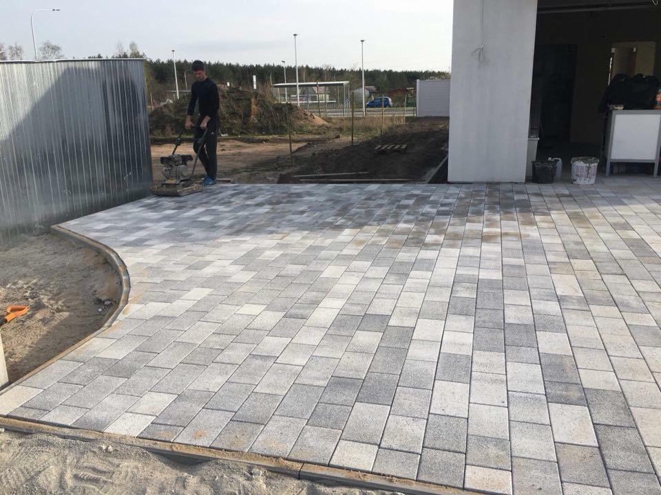 Prace brukarskie w toku: zagęszczarka ubija szaro-białą kostkę brukową ułożoną we wzór, widoczne obrzeża i przygotowane podłoże.