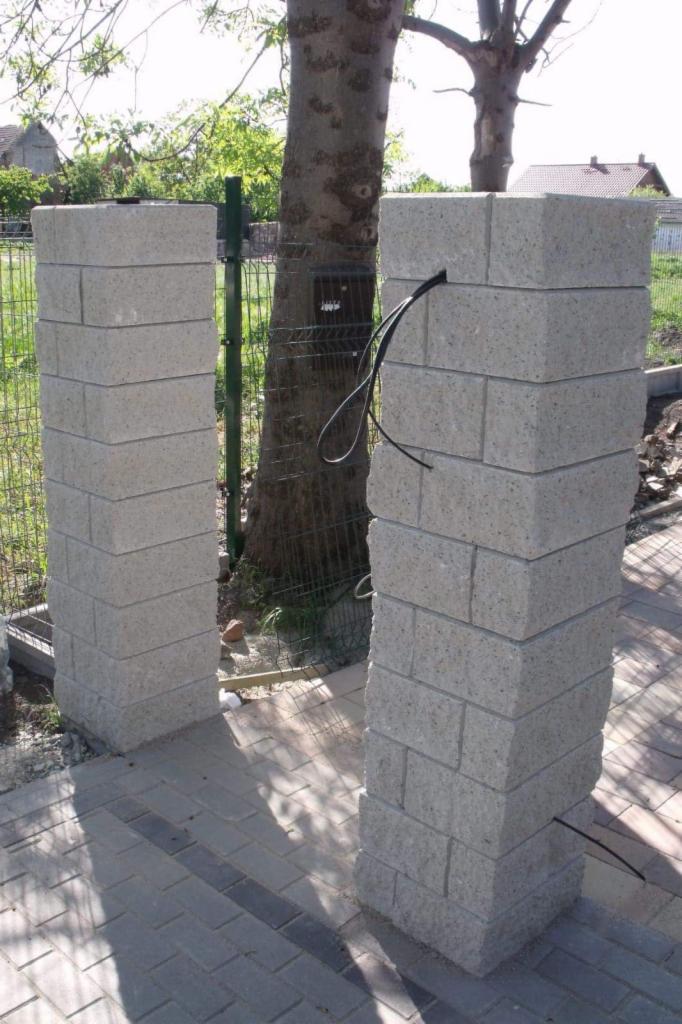 Dwa słupki z bloczków betonowych tworzące wejście, z widocznym okablowaniem w jednym słupku, otoczone kostką brukową i siatką ogrodzeniową, z drzewem w tle.