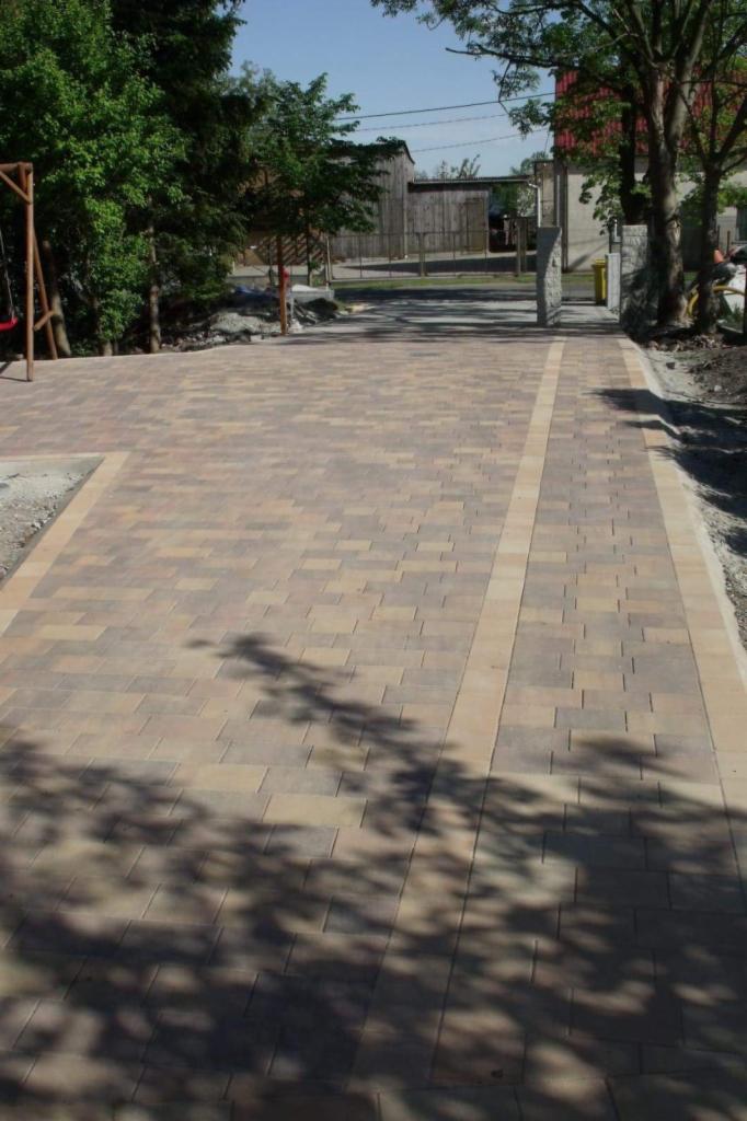 Ułożona dwukolorowa nawierzchnia z kostki, z widocznymi cieniami drzew, prowadząca w kierunku drewnianych budynków w tle.