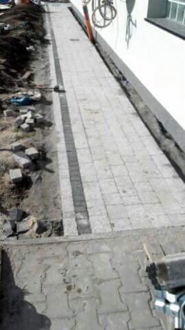 Ścieżka z jasnej kostki brukowej z ciemniejszym pasem pośrodku, w trakcie układania, widoczne narzędzia i nieułożone kostki przy krawędzi