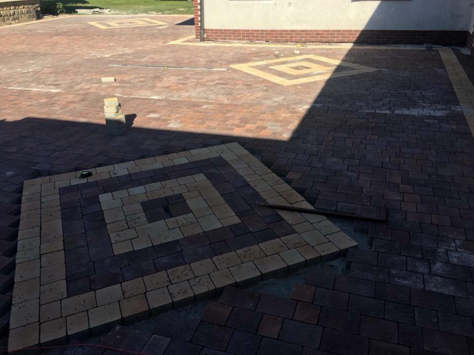 Ułożona część nawierzchni z kostki brukowej w geometryczny wzór, widoczne niedokończone fragmenty i narzędzia, w tle budynek z ceglanym cokołem i ułożona nawierzchnia z innym wzorem.
