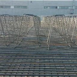 Siatka zbrojeniowa z wystającymi pionowymi elementami, przygotowana do zalania betonem, na tle hali przemysłowej.