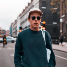 Młody mężczyzna w okularach przeciwsłonecznych i czapce z daszkiem, pozujący na ulicy w mieście z budynkami z cegły w tle, ubrany w granatową bluzę i białą torbę na ramię.