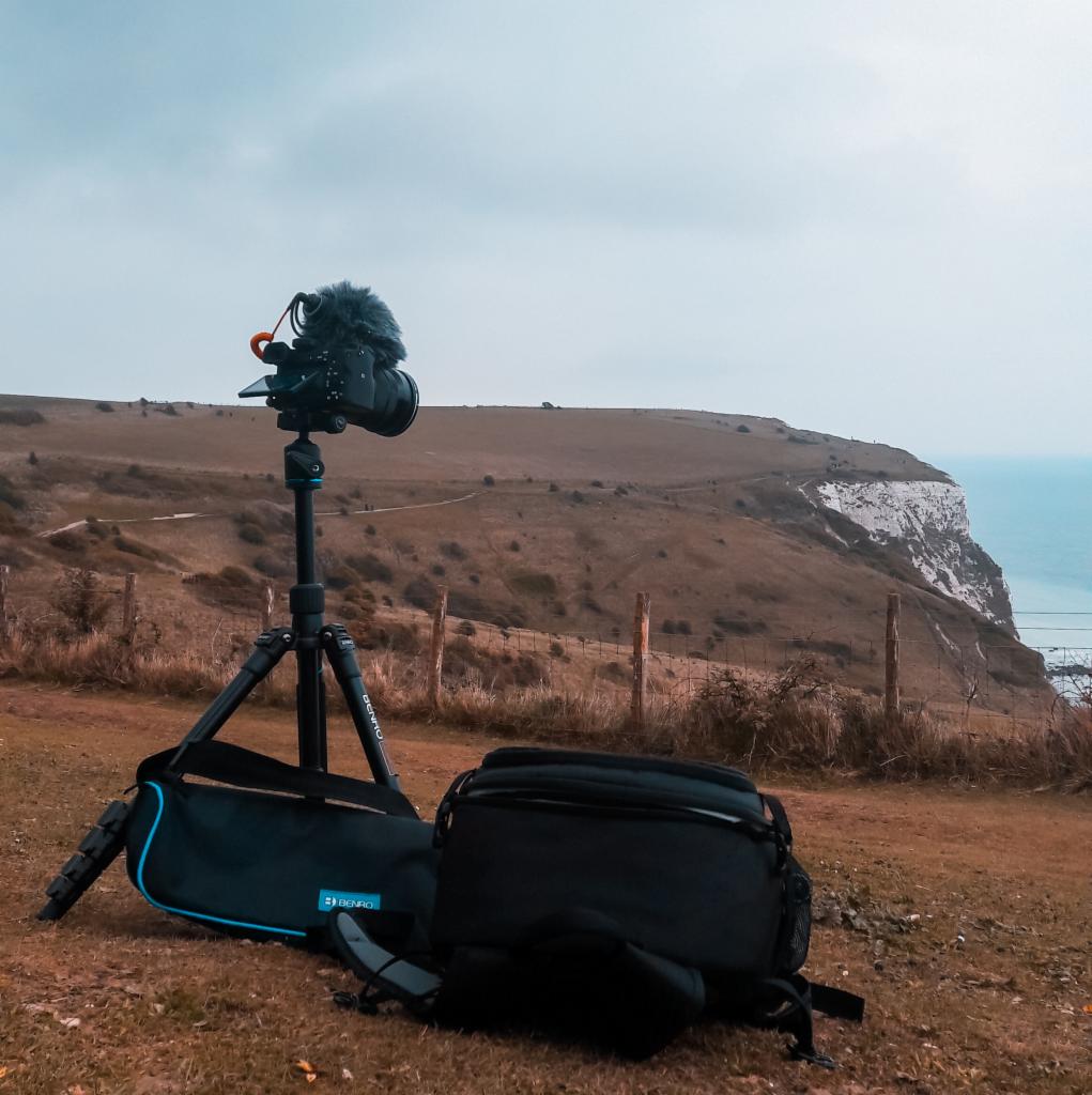 Aparat na statywie z mikrofonem typu 'deadcat' i torbami fotograficznymi na tle klifu i morza w pochmurny dzień.