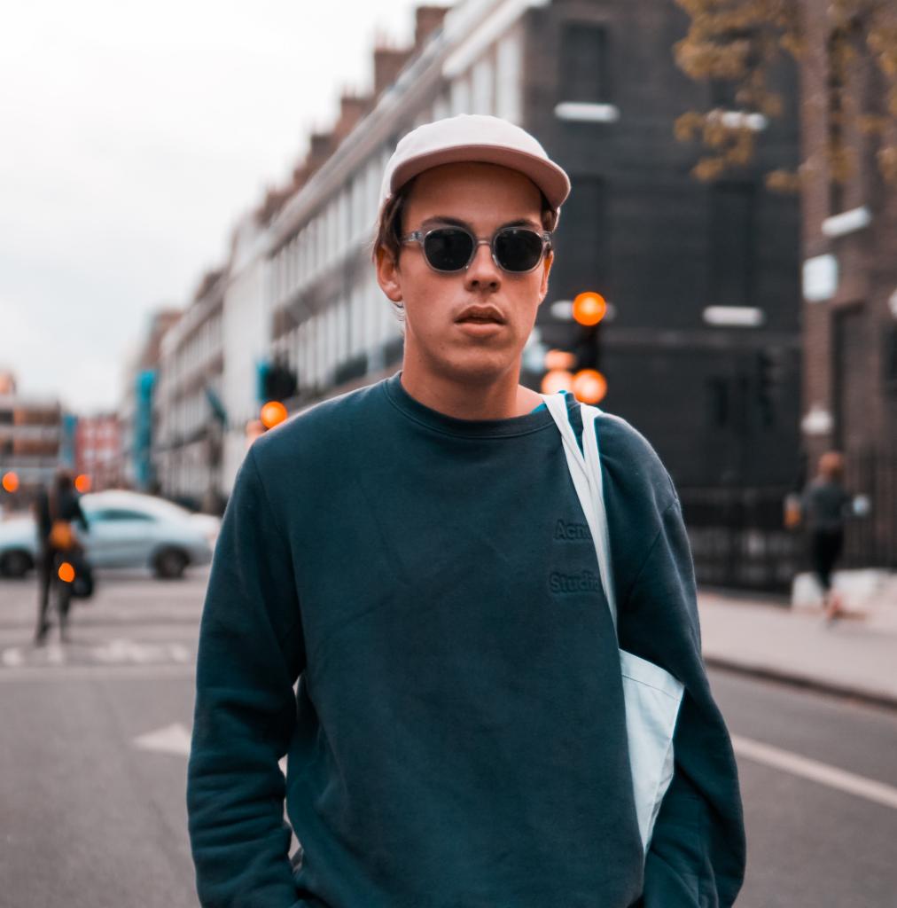 Młody mężczyzna w okularach przeciwsłonecznych i czapce z daszkiem, pozujący na ulicy w mieście z budynkami z cegły w tle, ubrany w granatową bluzę i białą torbę na ramię.