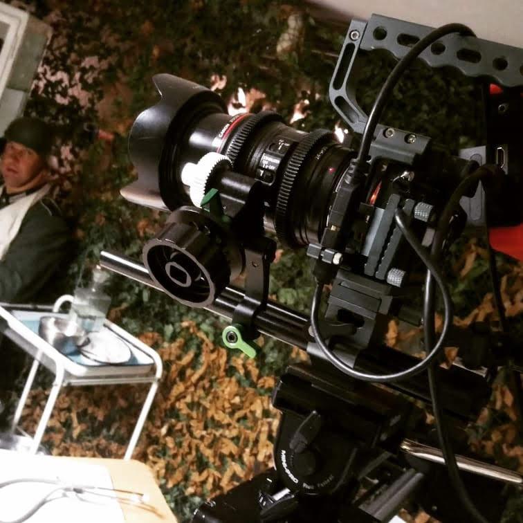 Profesjonalna kamera filmowa Canon na statywie z osprzętem, ustawiona na planie filmowym z aktorem w tle, przygotowującym się do sceny.