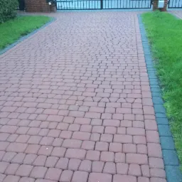 Czerwona kostka brukowa ułożona w zygzak na podjeździe, obrzeża z ciemnej kostki, trawnik po bokach, brama w tle.