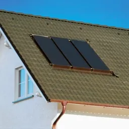 Trzy panele kolektorów słonecznych, zamontowane w dachu skośnym domu jednorodzinnego z białymi ścianami i brązową dachówką, widoczne rynny miedziane. Błękitne, bezchmurne niebo w tle.
