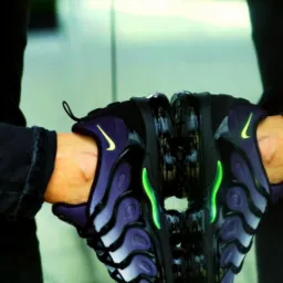 Dwie ręce trzymają parę fioletowo-czarnych butów sportowych Nike Air Max Plus TN, odbijających się w szklanej powierzchni.