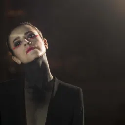 Portret kobiety w czarnej marynarce z artystycznym makijażem w odcieniach czerni i czerwieni na ciemnym tle.