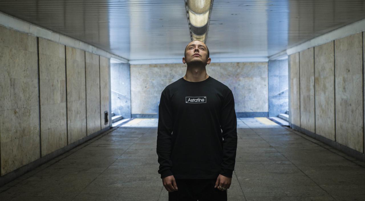 Mężczyzna w czarnej koszulce z długim rękawem i białym logo 'Astatine' spogląda w górę w betonowym przejściu podziemnym z oświetleniem sufitowym.
