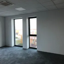 Puste, świeżo pomalowane biuro z trzema oknami w czarnych ramach, szarą wykładziną dywanową i podwieszanym sufitem z kratką.