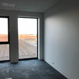 Puste, nowocześnie wykończone biuro z dwoma oknami z czarnymi ramami, szarą podłogą i białymi ścianami, widok na pole za oknem.