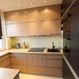 Nowoczesna kuchnia z beżowymi szafkami i ciemnobrązową zabudową piekarnika; widoczny czarny zlew, płyta indukcyjna, blat z delikatnym wzorem, roleta w oknie i butelka Jack Daniel's jako element...