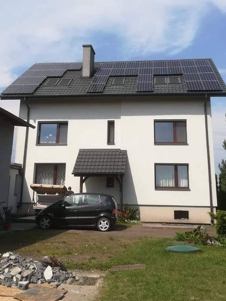 Dwupiętrowy dom z ciemną dachówką i panelami słonecznymi na dachu, na trawniku zaparkowany czarny samochód osobowy, w tle huśtawka ogrodowa.