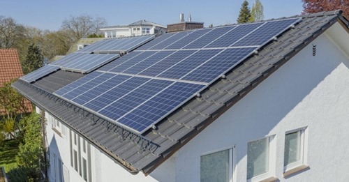 Panele fotowoltaiczne na dachu domu jednorodzinnego z jasną elewacją i ciemną dachówką, widoczne z perspektywy lekko z góry.