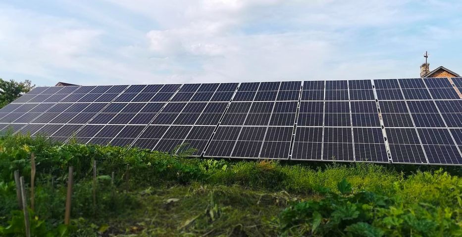 Rząd paneli słonecznych zamontowanych na dachu budynku, widocznych z perspektywy ogrodu z bujną roślinnością, pod błękitnym niebem.