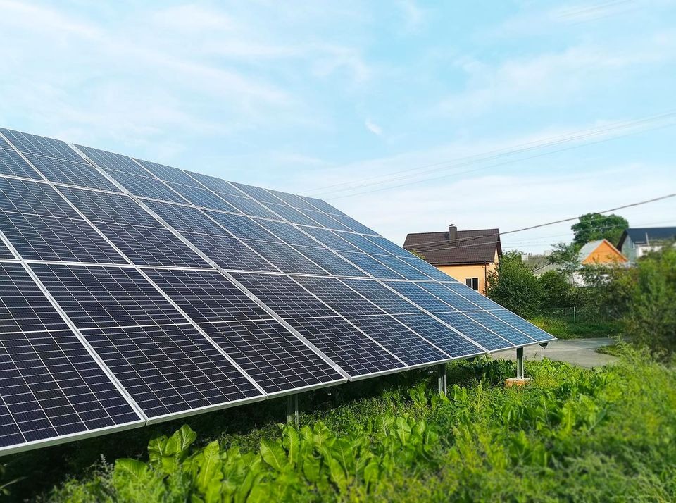 Panel słoneczny ustawiony pod kątem na tle zieleni i domów, widoczne przewody elektryczne na niebie.