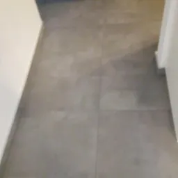 Szare, kwadratowe płytki podłogowe ułożone w korytarzu, widoczne fugi, ściany pomalowane na biało, w tle ciemna podłoga w innym pomieszczeniu.