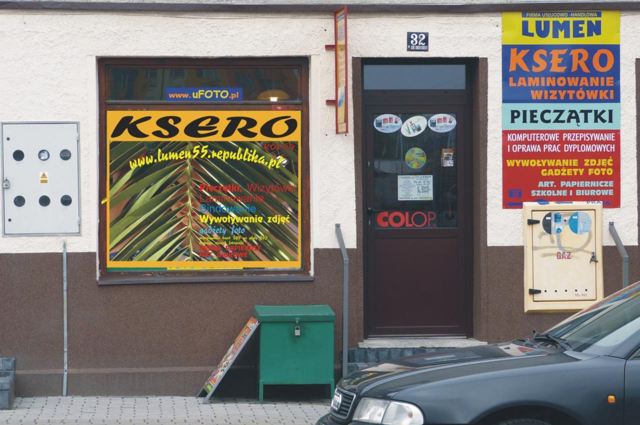 Fasada punktu usługowego z szyldami 'Ksero Lumen', 'Colop', oklejone okno z ofertą usług: pieczątki, wizytówki, laminowanie, bindowanie, wywoływanie zdjęć, gadżety foto, na zewnątrz skrzynka gazowa...