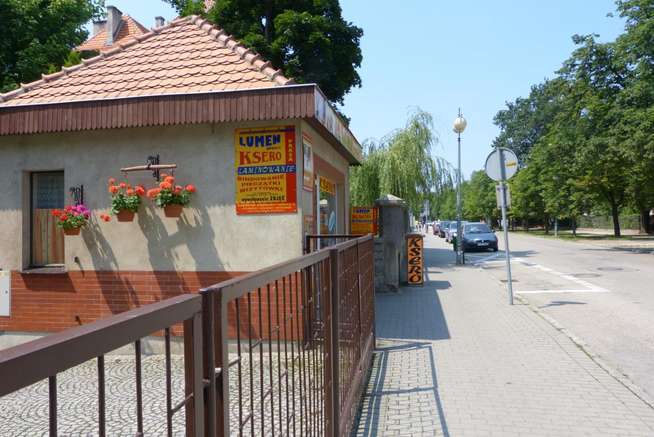 Budynek punktu ksero z reklamami na ścianie, wiszącymi doniczkami z kwiatami i brązowym ogrodzeniem, widok z chodnika w słoneczny dzień.