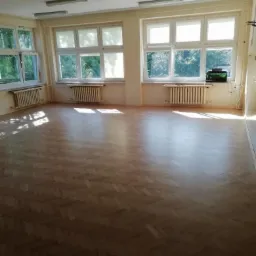 Jasne, przestronne wnętrze z parkietem ułożonym w jodełkę, oświetlone naturalnym światłem wpadającym przez okna. Widoczne grzejniki i akwarium pod oknem.