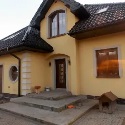 Żółty dom jednorodzinny z brązowymi oknami i drzwiami, czarnym dachem z dachówką, widoczne schody wejściowe, garaż z okrągłym oknem, buda dla psa i mały stołek przed wejściem.