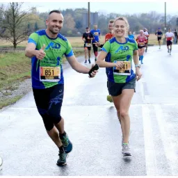 Uśmiechnięty mężczyzna z podniesionym kciukiem i kobieta biegną ramię w ramię podczas biegu ulicznego z numerami startowymi, mokra nawierzchnia, w tle inni biegacze i drzewa.