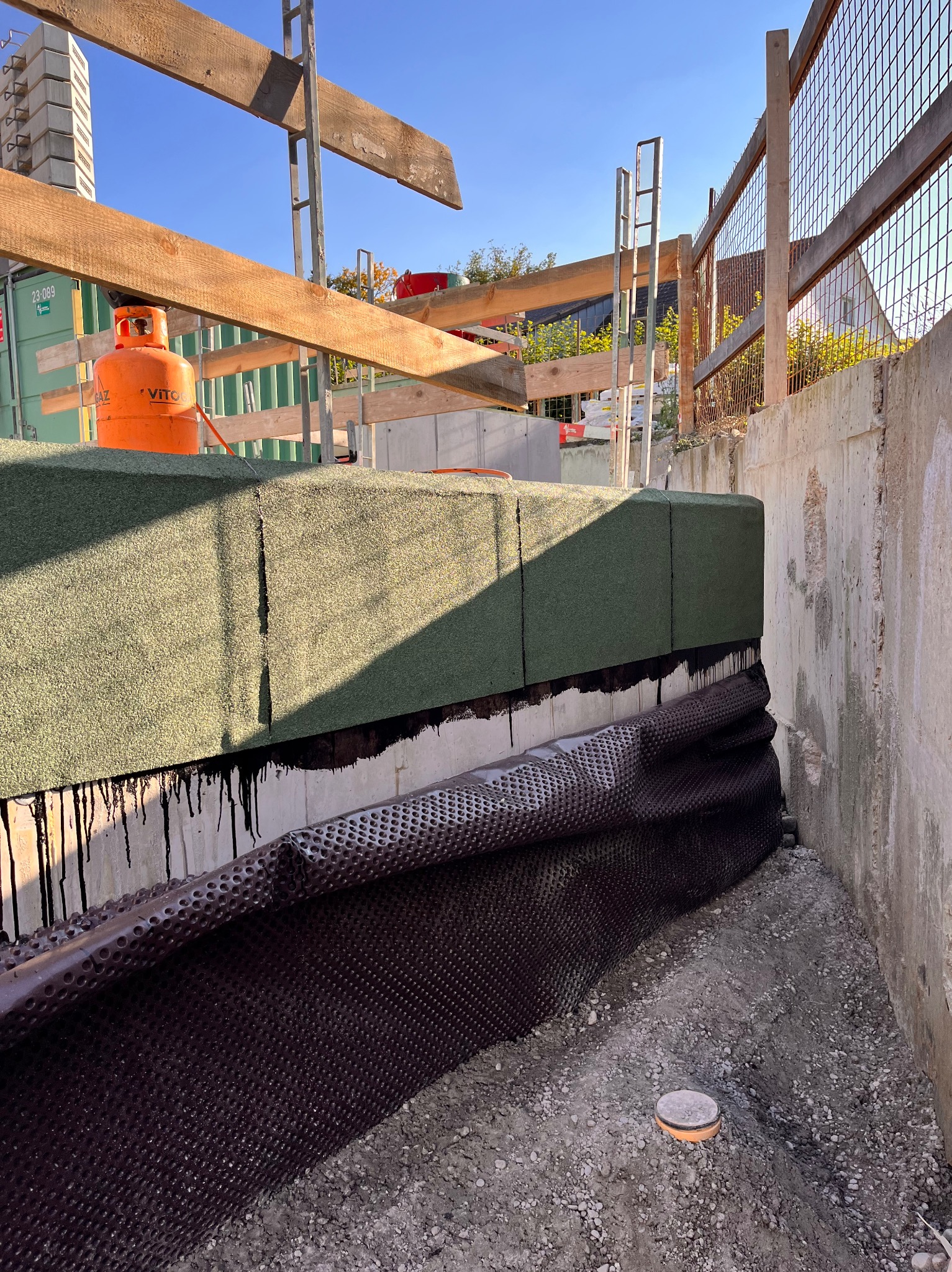 Izolacja fundamentów budynku w trakcie prac. Widoczna membrana kubełkowa, izolacja termiczna i hydroizolacja. W tle butla gazowa i elementy konstrukcji.