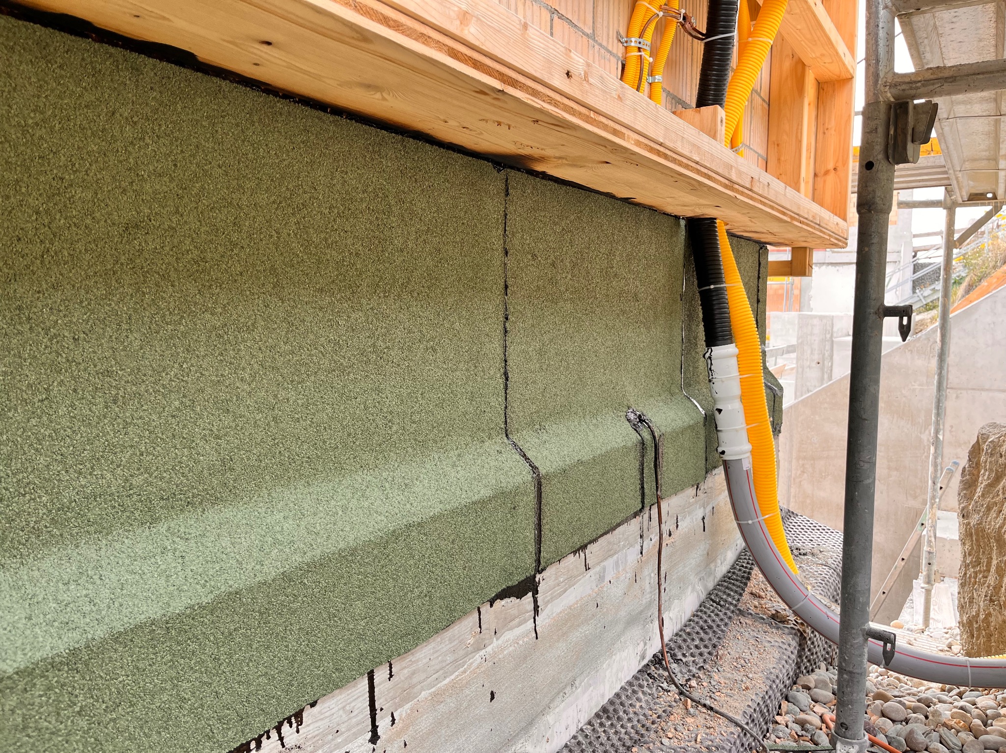 Ocieplenie ściany budynku z widocznymi rurami instalacyjnymi w żółtej osłonie, zamocowanymi do drewnianej konstrukcji. Widoczne detale izolacji i elementy rusztowania.