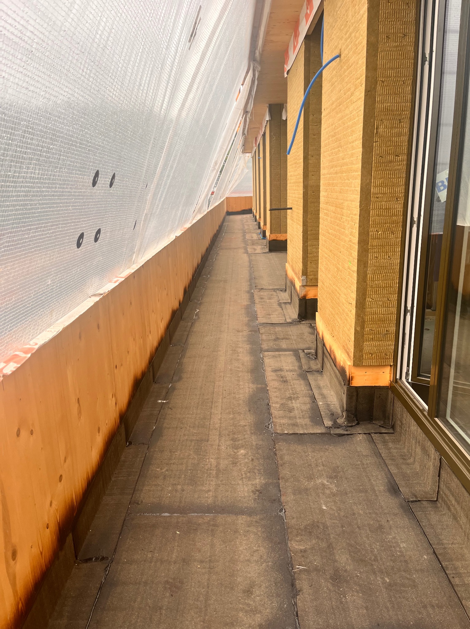 Dach pokryty papą termozgrzewalną, zabezpieczony folią ochronną. Widoczne ocieplenie ścian budynku wełną mineralną. Prace dekarskie w toku, detale wykończeniowe.