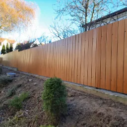 Nowoczesne ogrodzenie panelowe z pionowych desek w kolorze jasnego drewna, zamontowane na betonowej podmurówce, widoczne na tle ogrodu z drzewami i krzewami.