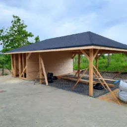 Drewniana konstrukcja z czarnym dachem w trakcie budowy, widoczne worki z materiałami budowlanymi i ułożona kostka brukowa w tle krajobraz z zielenią.