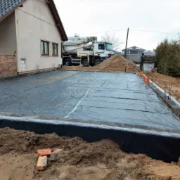 Przygotowanie podłoża pod wylewkę fundamentów: czarna folia izolacyjna na podsypce, widoczny betonowy obrys z wystającymi prętami zbrojeniowymi, w tle betoniarka i pryzma piasku.