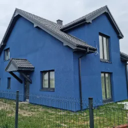 Nowo ocieplony dom jednorodzinny z niebieską elewacją, grafitowym dachem i stolarką okienną, widoczny zza metalowego ogrodzenia panelowego.