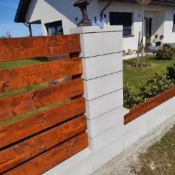 Nowoczesne ogrodzenie z drewnianymi przęsłami w kolorze brązu i szarymi słupkami z betonowych bloczków; w tle widoczny dom z ogrodem, na słupku leży wkrętarka akumulatorowa.