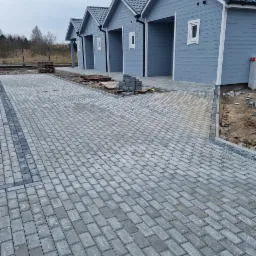 Ułożona szara kostka brukowa z ciemniejszym obramowaniem, prowadząca do szeregu garaży o niebieskich ścianach i białych obramowaniach okien, na obrzeżach widoczne palety i przygotowane kostki.