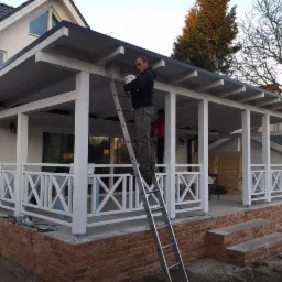 Mężczyzna maluje białą altanę ogrodową z drabiny, widoczne ceglane podmurówki i elementy drewniane.