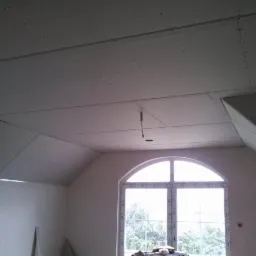 Poddasze z oknem łukowym w trakcie ocieplania, widoczne płyty gipsowo-kartonowe na skosach i suficie, przygotowane do szpachlowania, zwisający kabel elektryczny.