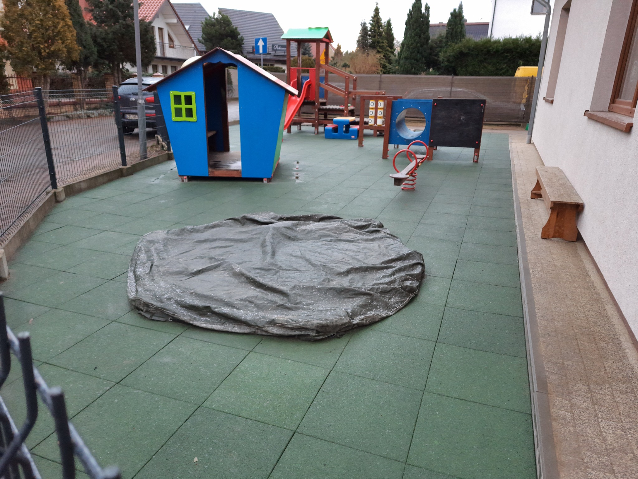 Plac zabaw z elementami dla dzieci, w tym domek, zjeżdżalnia i huśtawka, częściowo przykryty plandeką, wyłożony zieloną gumową nawierzchnią.