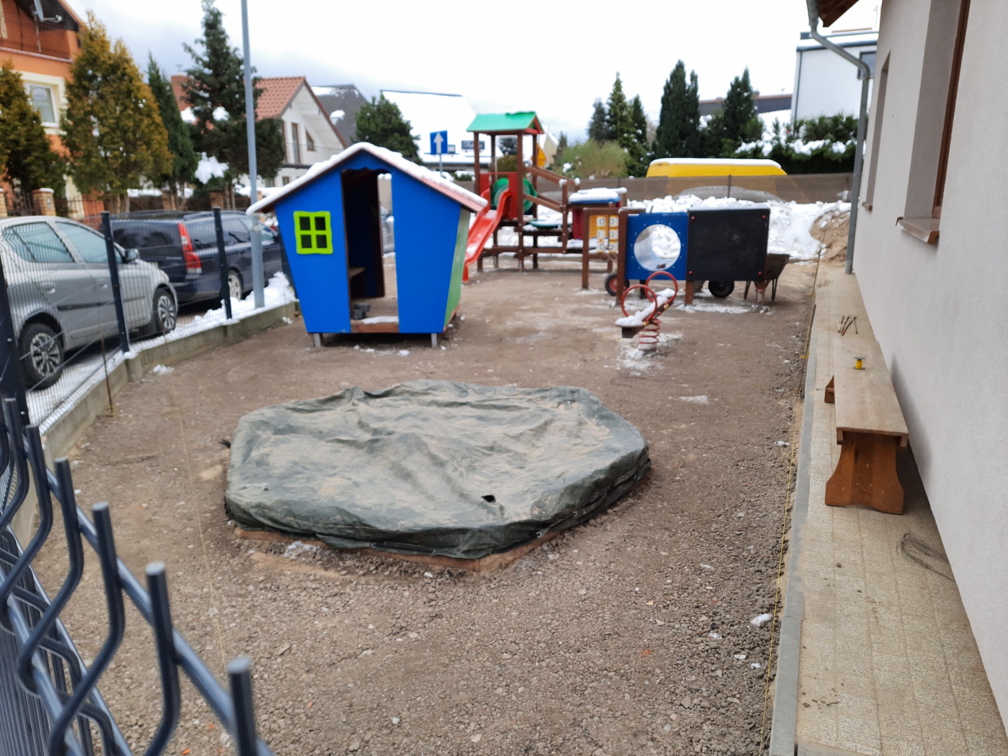 Plac zabaw w trakcie budowy z niebieskim domkiem, huśtawką i piaskownicą przykrytą plandeką, widok zza ogrodzenia.