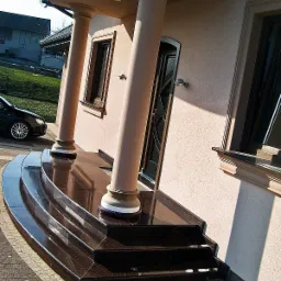 Eleganckie, ciemne, polerowane schody wejściowe z kamienia, prowadzące do domu z kolumnami. Widoczny fragment elewacji, okna i samochód w tle.