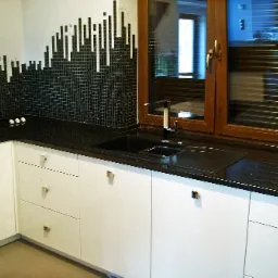 Kuchenny blat z czarnego kamienia z wbudowanym zlewem, mozaika na ścianie imitująca skyline miasta, białe szafki z kwadratowymi uchwytami, drewniane okno z żaluzjami.