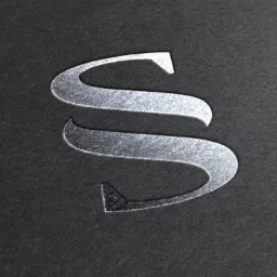 Srebrna litera S wytłoczona na ciemnoszarym, teksturowanym papierze, widoczna faktura papieru.