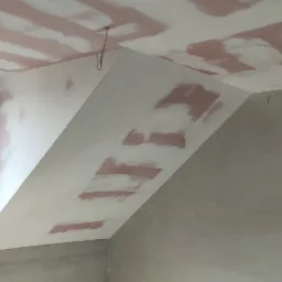 Skośny sufit poddasza w trakcie prac przygotowawczych, widoczne fragmenty nałożonej zaprawy wyrównującej na płytach kartonowo-gipsowych i suficie, szary odcień ścian w tle.