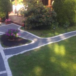 Radbruk - Ogród z ścieżką z kostki brukowej w kształcie litery Y, obrzeża z jasnego kamienia, rabata z kwiatami, kobieta w piżamie i klapkach, trawnik, tuje i lampki solarne.